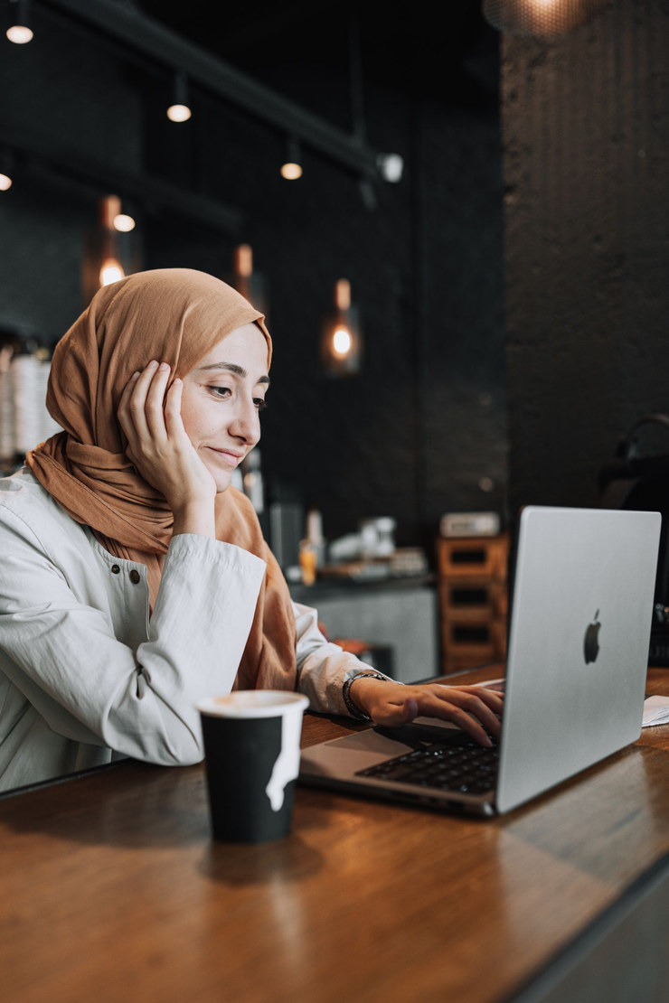 A woman in a headscarf lingering over her laptop and coffee in a dark café.
